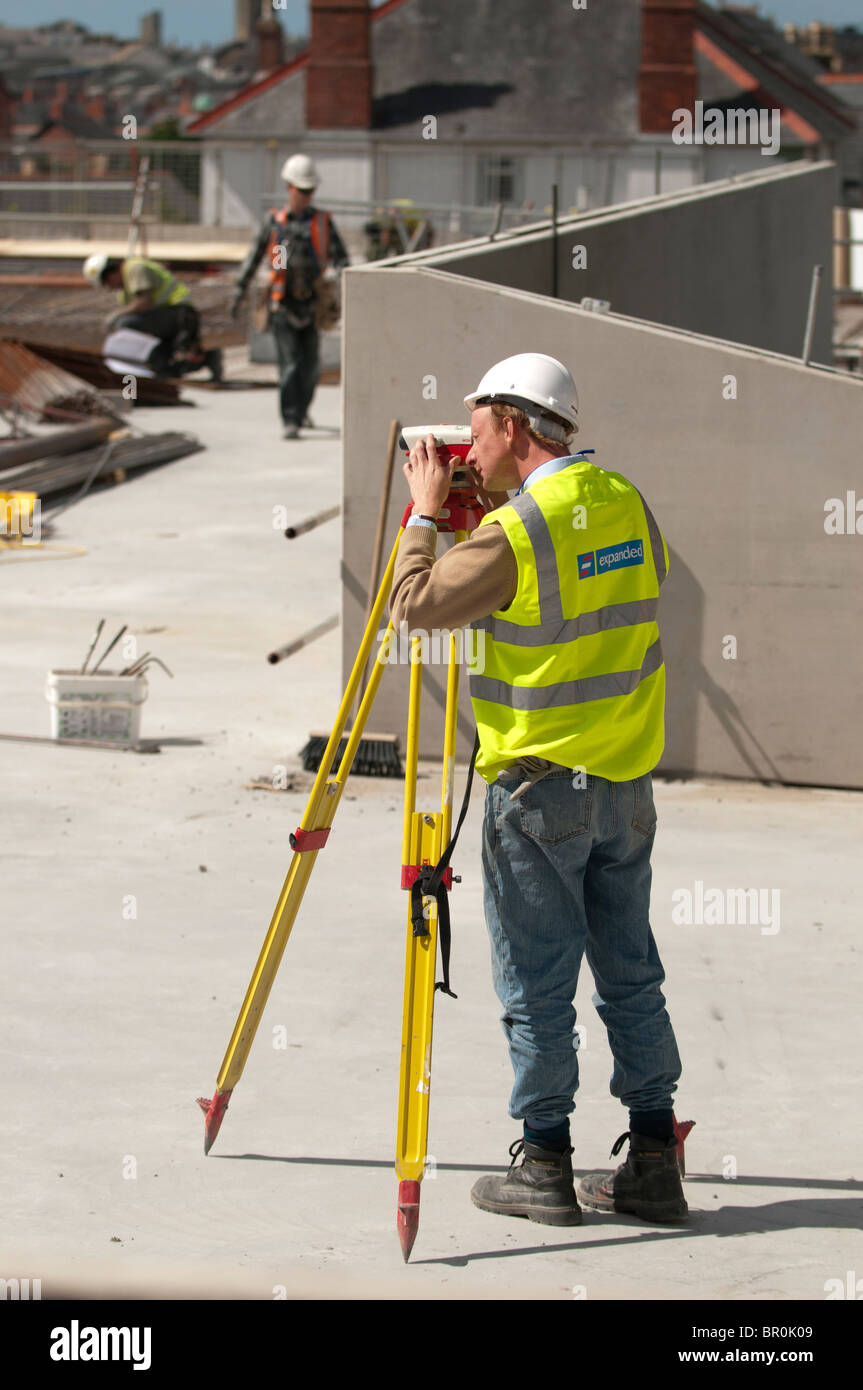 Un hombre, vistiendo chaqueta hi vis y completo equipo de EPI, trabajando en un sitio de construcción topografía la ubicación con una Leica teodolito, REINO UNIDO Foto de stock