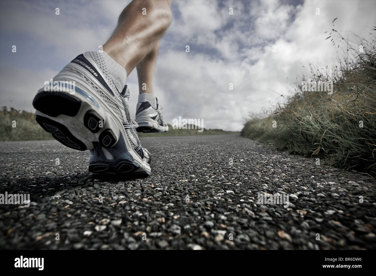 Una estrecha, ángulo de visión baja de un atleta masculino discurre por un camino asfaltado, con sus grandes músculos de la pierna mostrando Foto de stock