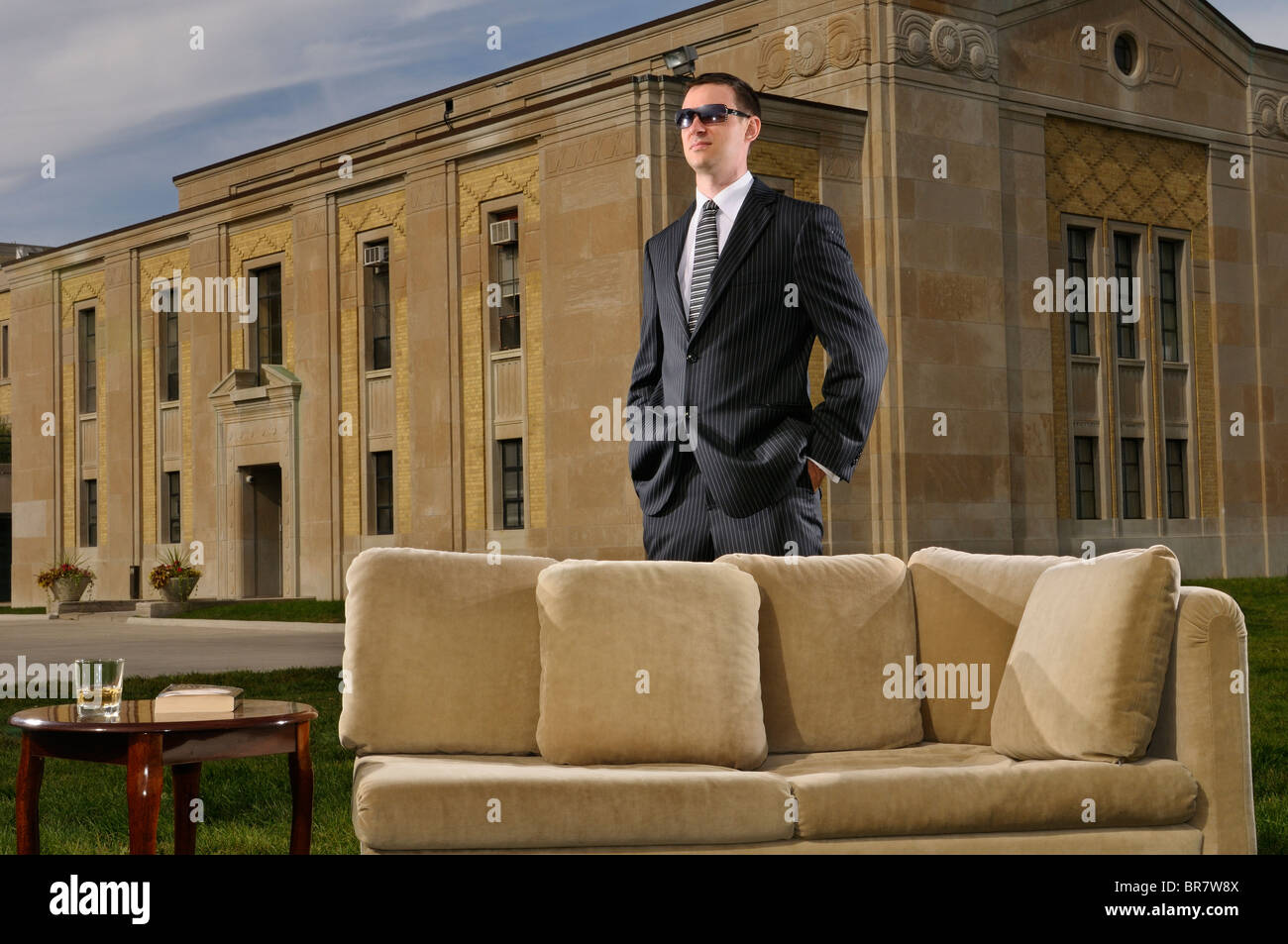 Joven rico en traje de pie detrás de un sofá y mesa en el césped en una mansión art deco Foto de stock