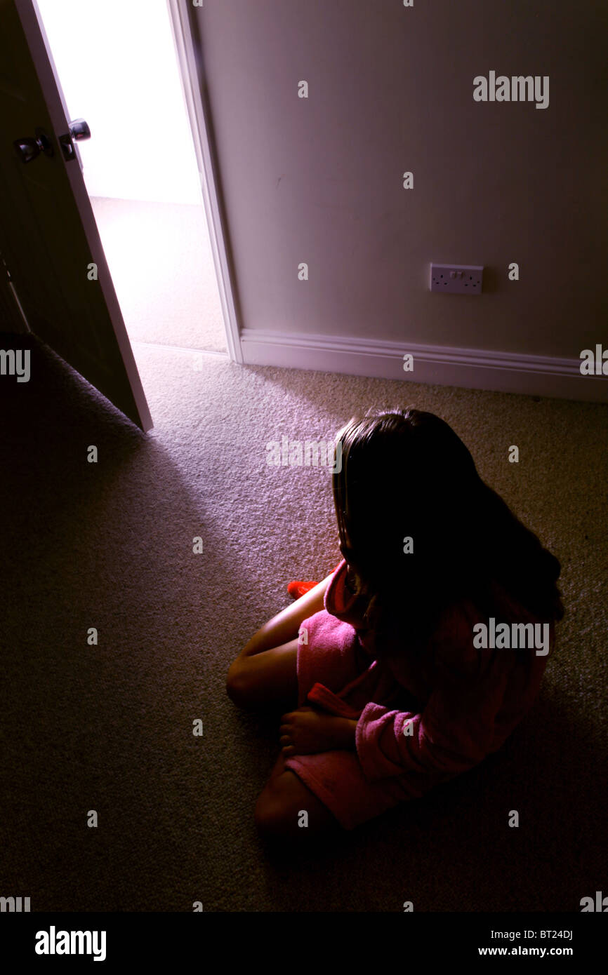 Chica sentada en una habitación oscura, vista posterior mirando a una puerta abierta, donde la luz se desliza. Foto de stock