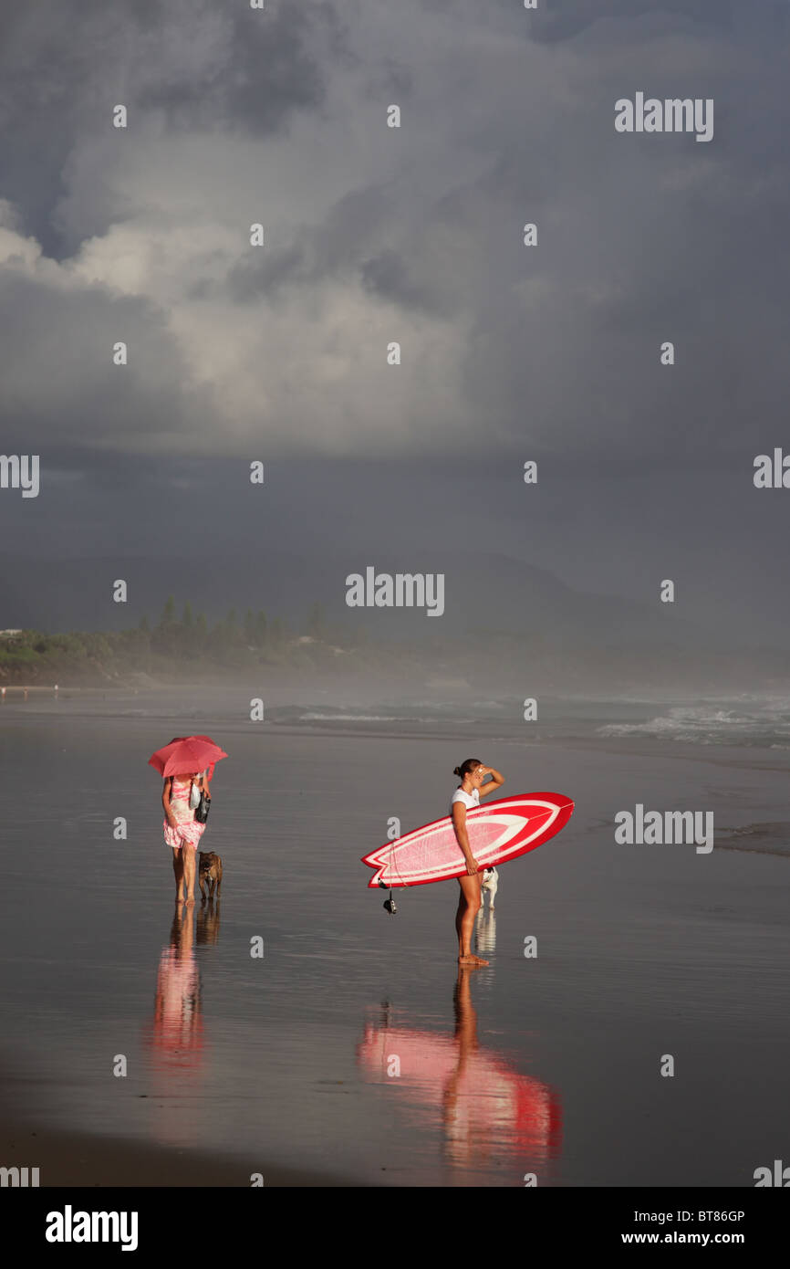 Niña con tabla de surf roja mirando al mar temprano en la mañana en la playa de Byron Bay, Australia. Foto de stock
