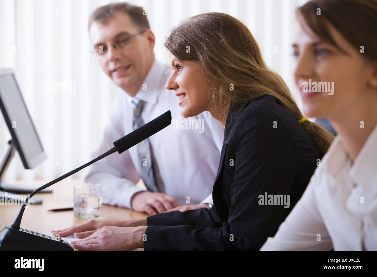 Mujer joven con éxito hablando en público el micrófono sentado a la mesa en negro cómodas sillas entre sus colegas Foto de stock
