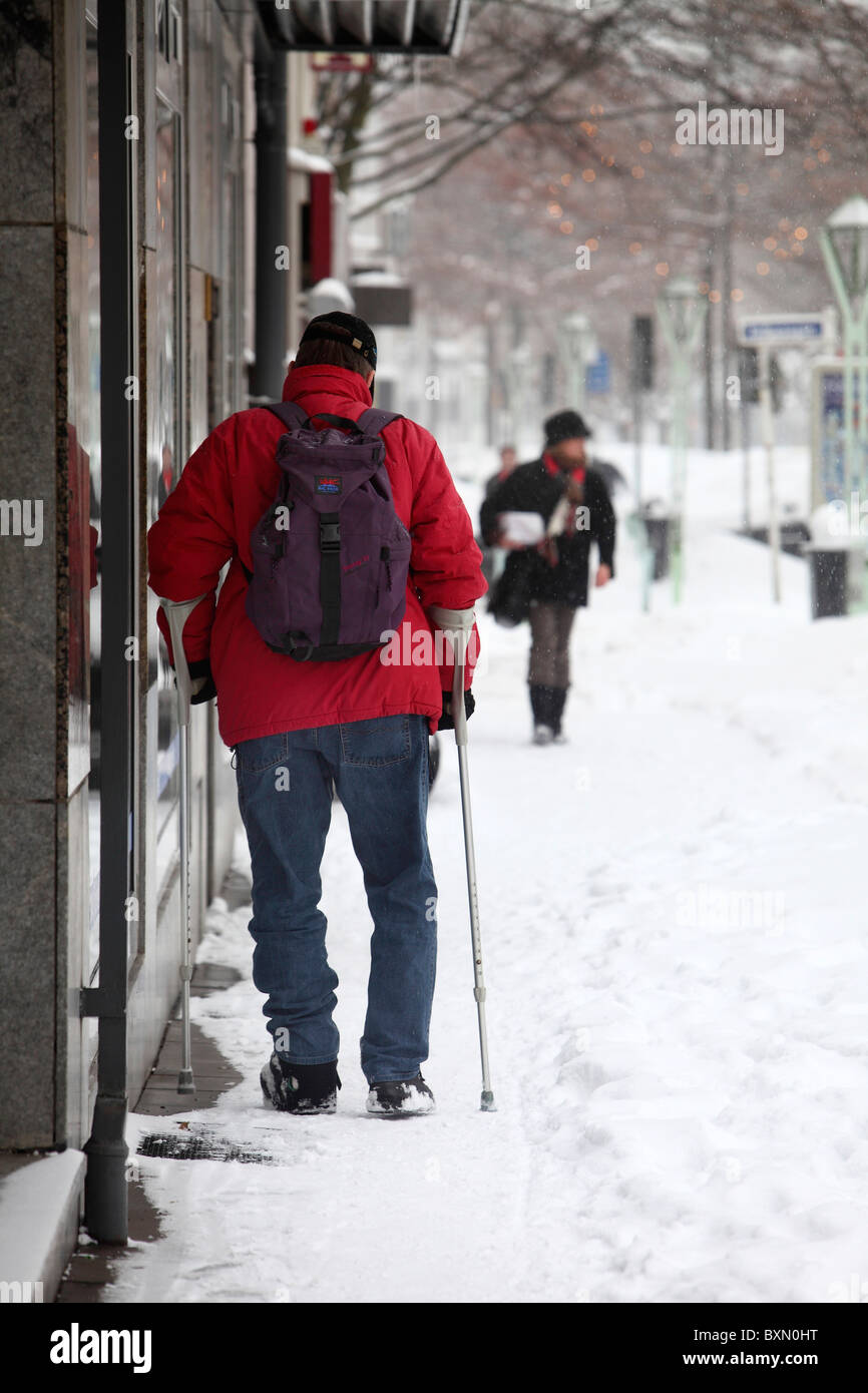 Hombre de edad avanzada con una muleta, intenta hacer su camino en un helado y cubierto de nieve en un camino peatonal de la ciudad. Foto de stock