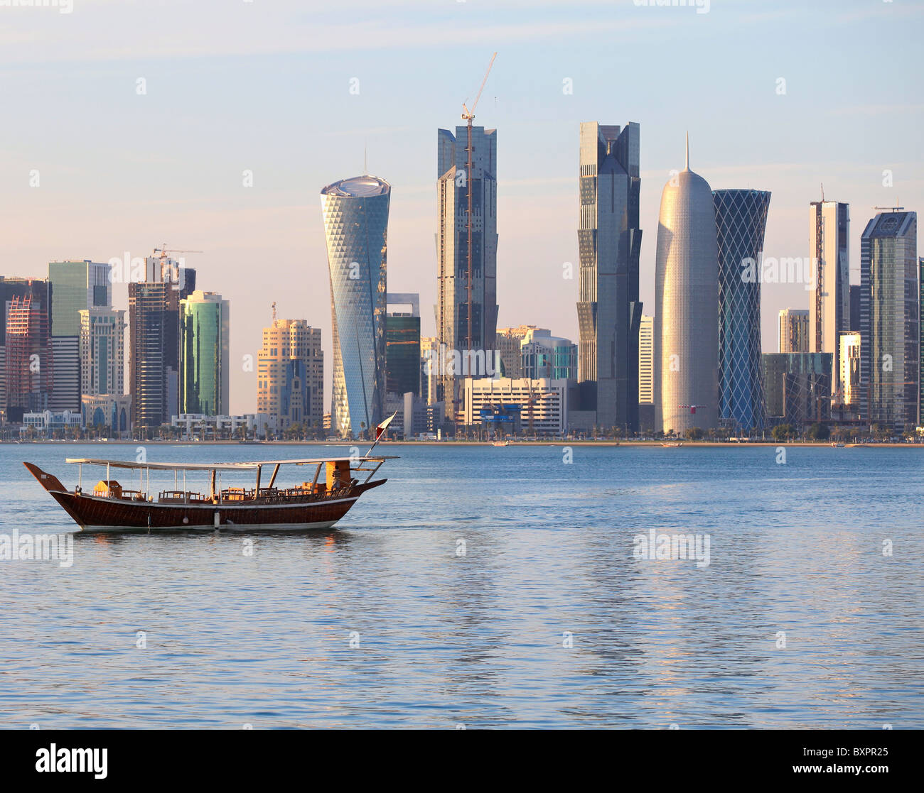 Un dhow regresa a puerto en Doha, Qatar, al atardecer, con modernos rascacielos de la ciudad en el fondo. Foto de stock