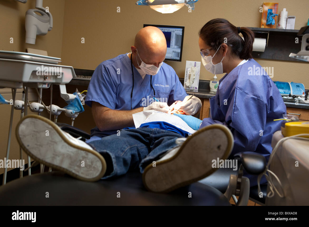 Anglo dentista trabaja en pacientes masculinos, mientras que entre las mujeres la asistente dental busca en la sala de tratamiento de clínica dental en Austin TX USA Foto de stock