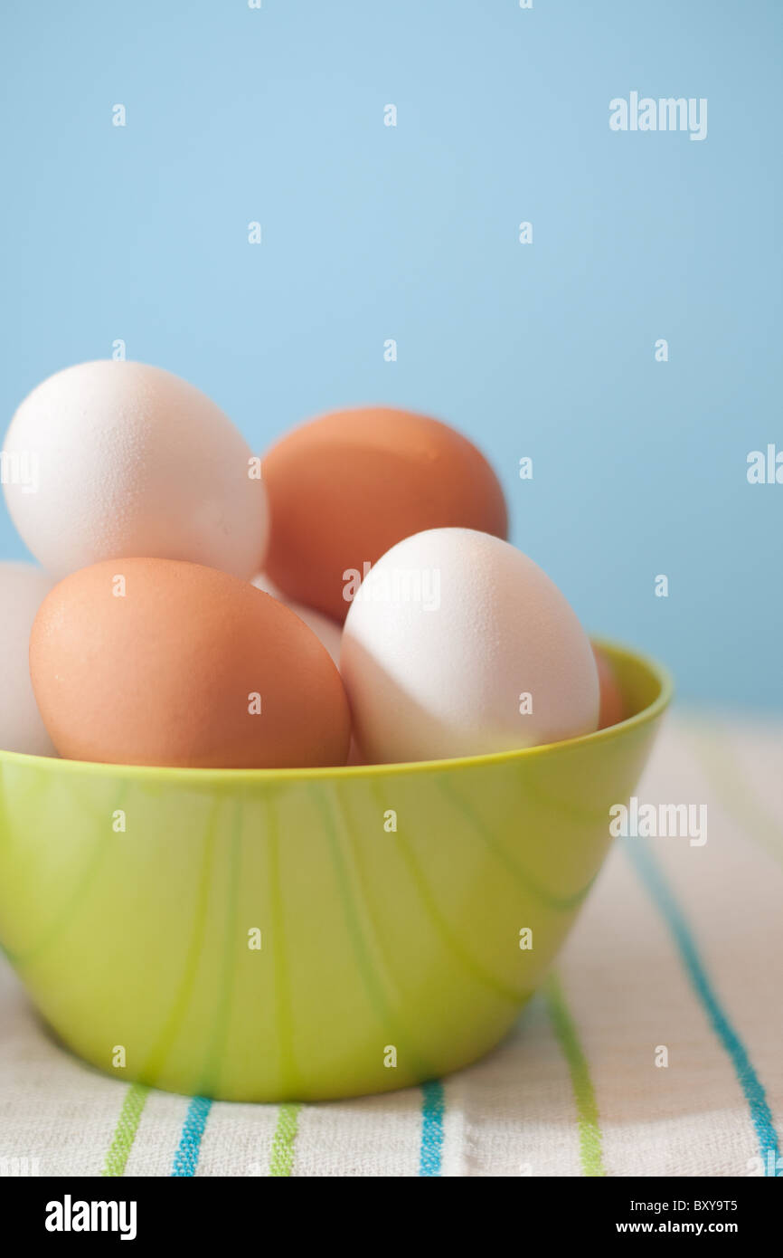 Marrón y blanco de los huevos de pollo en un tazón de rayas verdes sobre un paño de cocina con un fondo azul. Foto de stock
