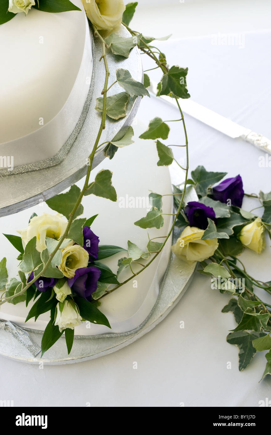 Pastel de boda blanco dos niveles cubiertos de flores púrpura y crema con ivy Foto de stock