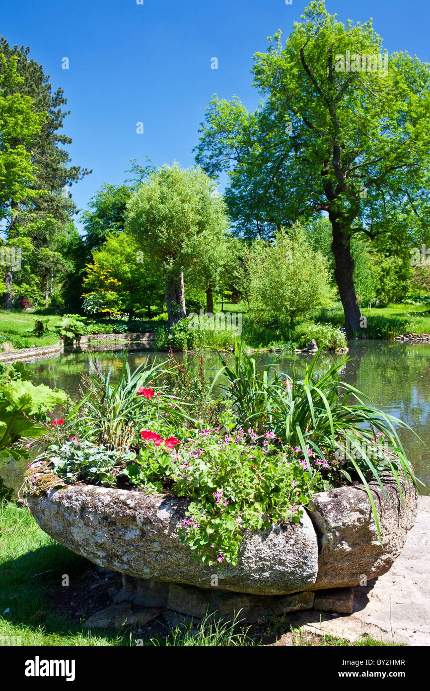 Una piedra sembradora con trailing geranios delante de un gran estanque ornamental o pequeño lago en un país inglés jardín en verano Foto de stock