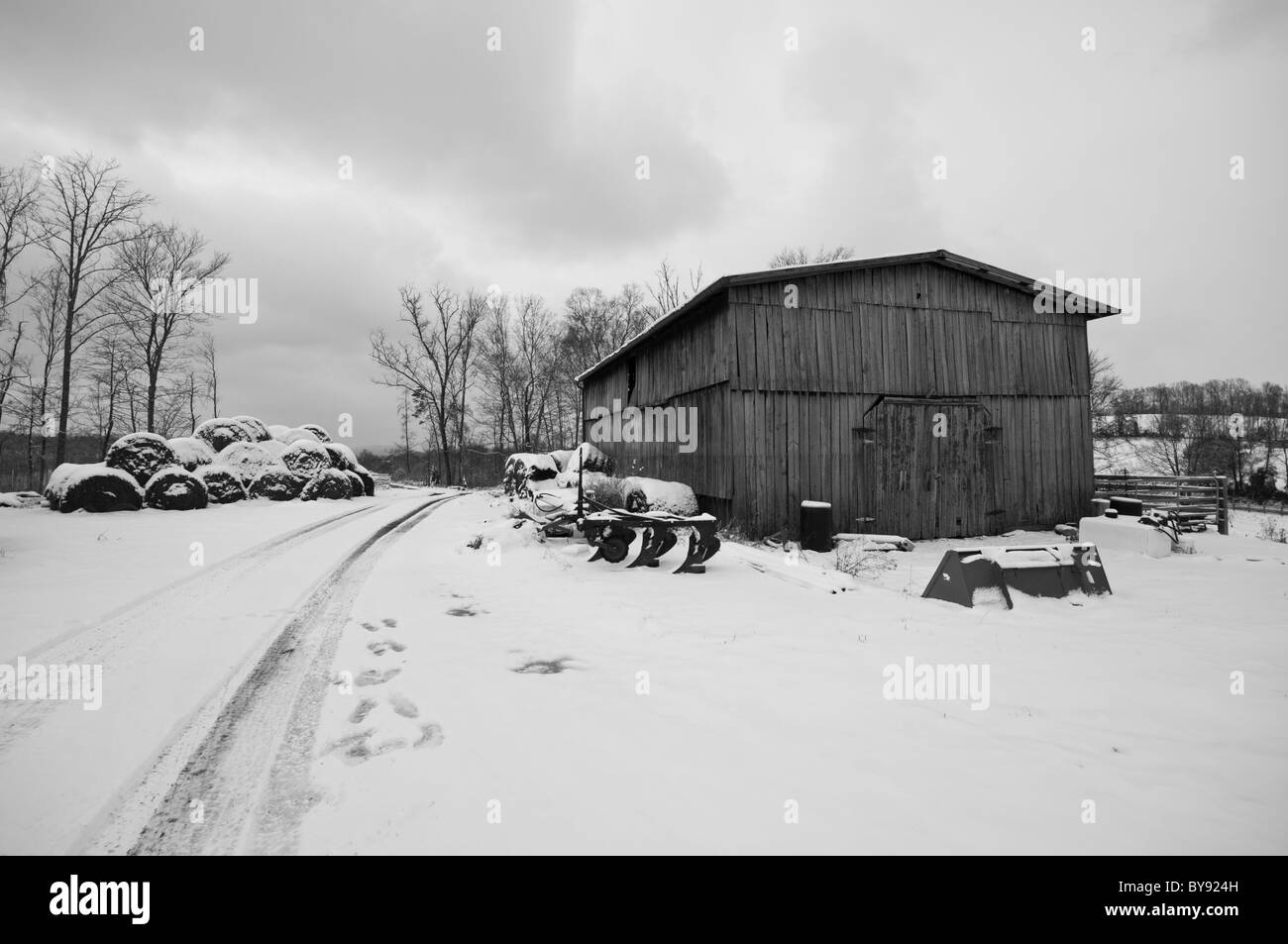 Escena de Invierno de un viejo granero junto a un Farm Road, ambas cubiertas de nieve. Un arado y otras herramientas del comercio son visibles. Foto de stock