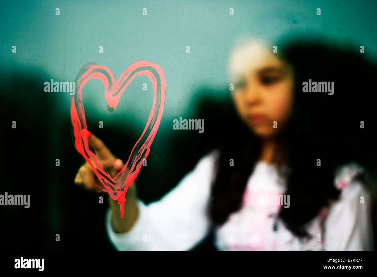 Chica dibuja con forma de corazón en la ventana sucia con su dedo en pintura roja. Foto de stock