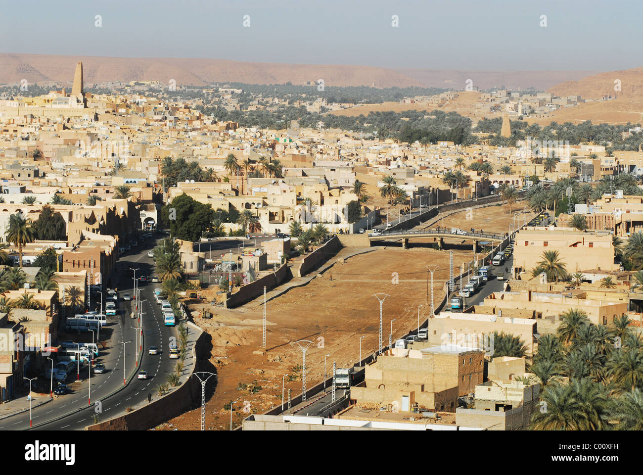 Ghardaia, Argelia, vista de la ciudad con los vehículos en carretera, vivienda de ladrillos de barro y alto minarete contra el cielo claro Foto de stock