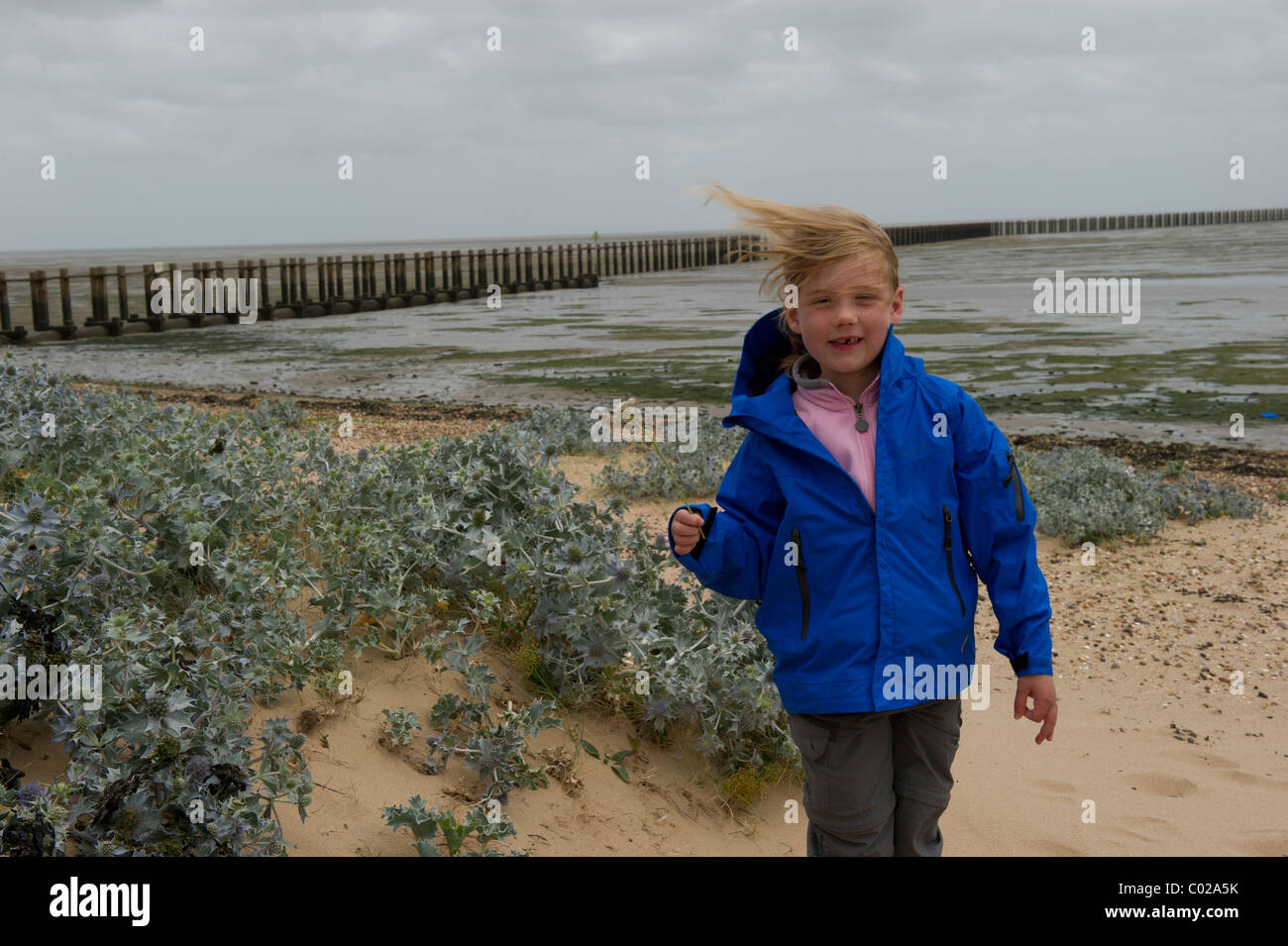 Joven chica blanca de pie en la playa muy ventosa mirando a la cámara y sonriendo. Guerra Mundial 2 de defensa del mar de fondo. Foto de stock