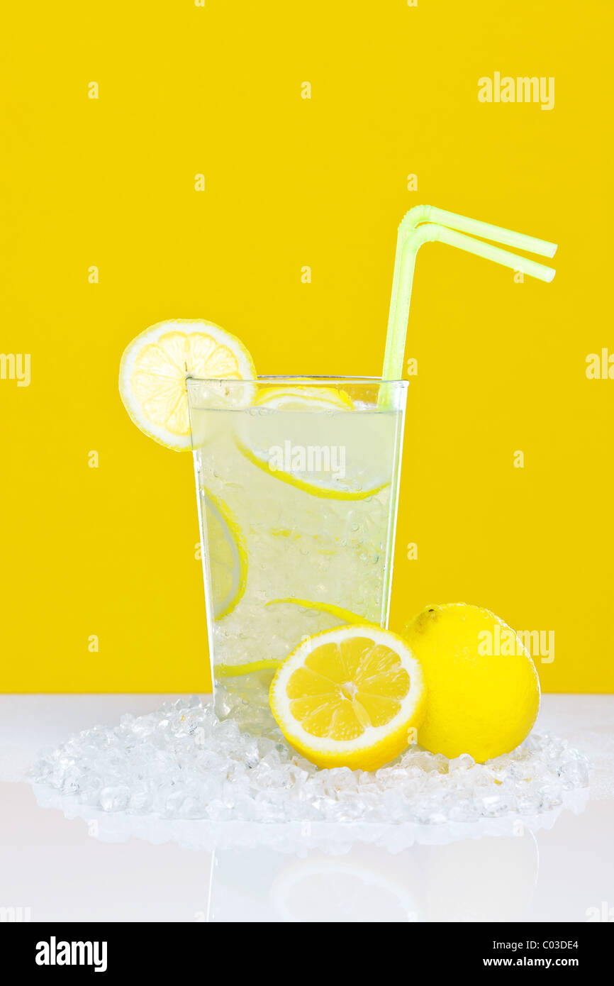 Foto de limonada tradicional en un vaso con hielo picado y rodajas de limón, en un cuadro blanco con fondo amarillo. Foto de stock