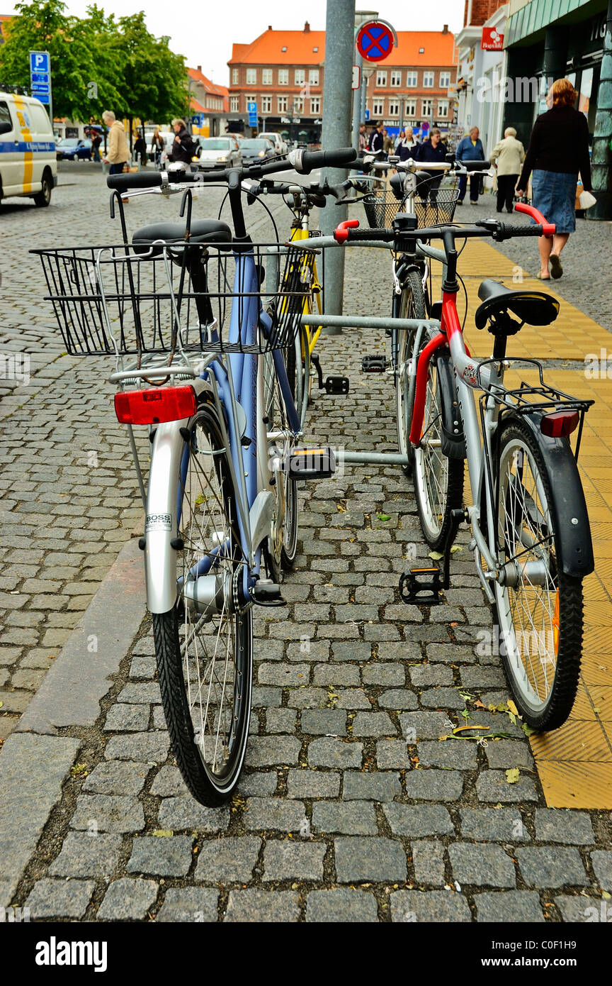 Bicicletas deja en un soporte de la bici oficial en una calle de adoquines en un concurrido centro comercial de Ronne Bornholm Foto de stock