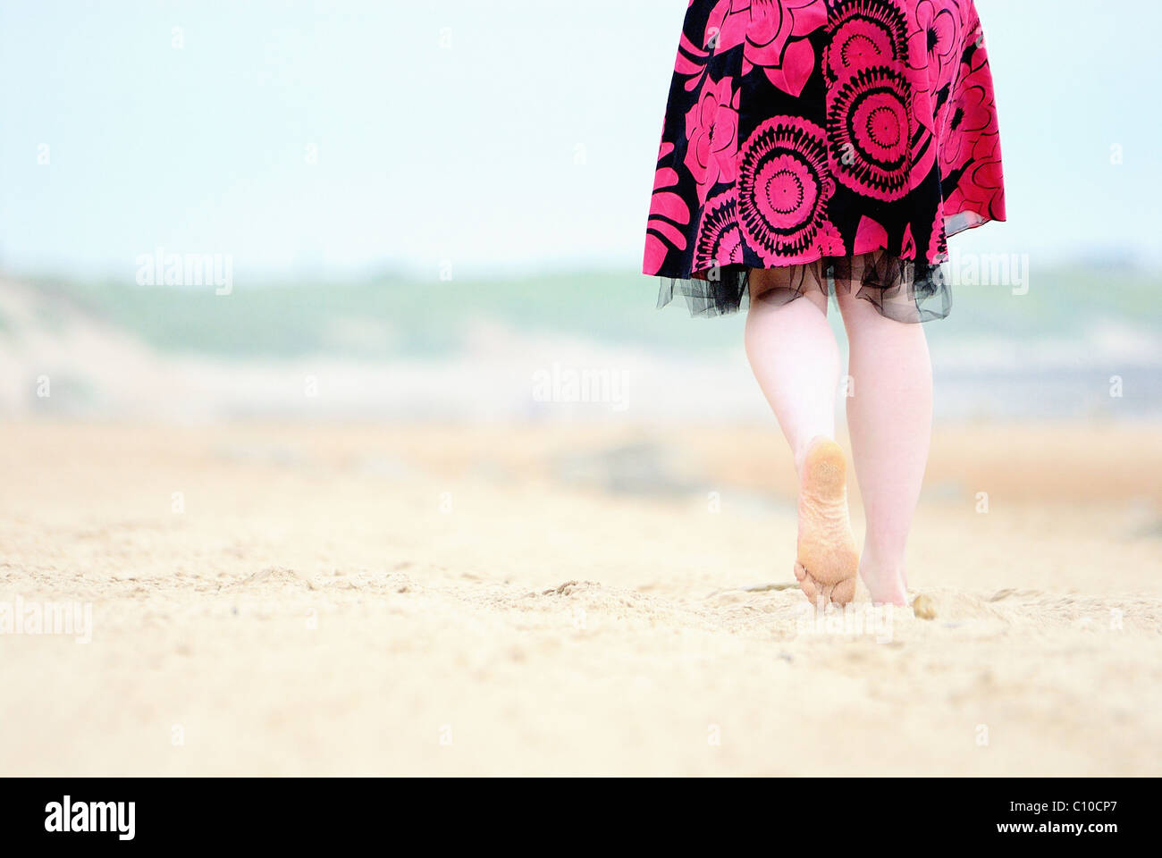 Un par de pies caminando por la playa en un vestido rosado y negro, caminando sobre la arena puedes ver la suela de un pie. Foto de stock