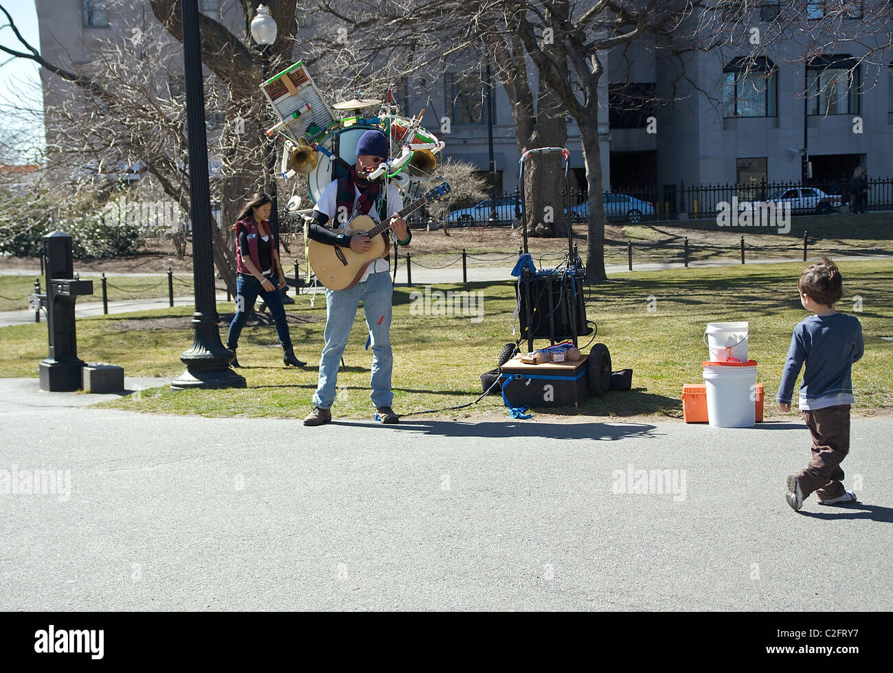 Un muchacho caminando a través de un parque se convierte a mirar hacia un hombre con una guitarra y realizando numerosos instrumentos atados a la espalda Foto de stock
