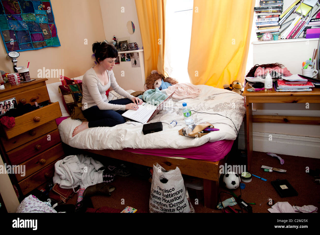 Una joven estudiante universitaria en el Reino Unido trabajan en su computadora portátil en la cama en un dormitorio desordenado Foto de stock