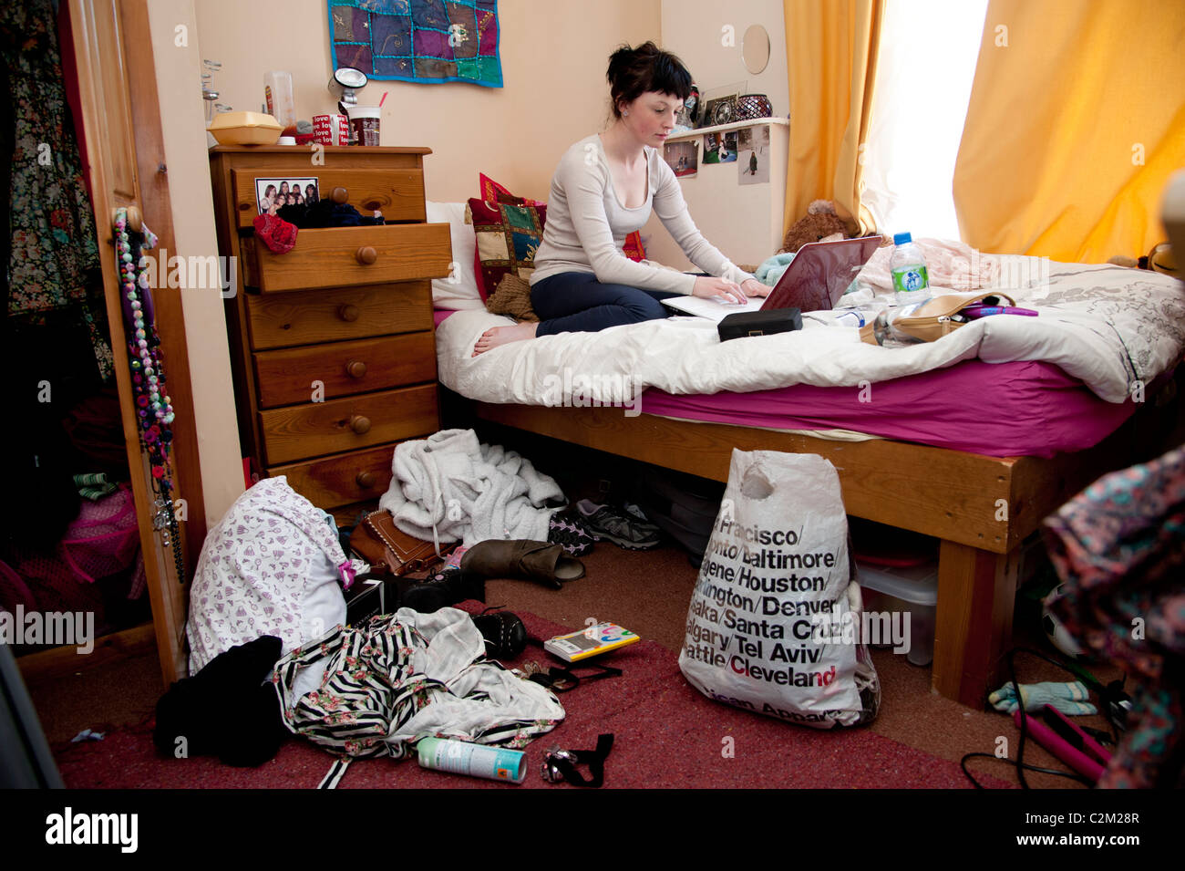 Una joven estudiante de la universidad de aberystwyth en el Reino Unido trabajan en su computadora portátil en la cama en un dormitorio desordenado Foto de stock