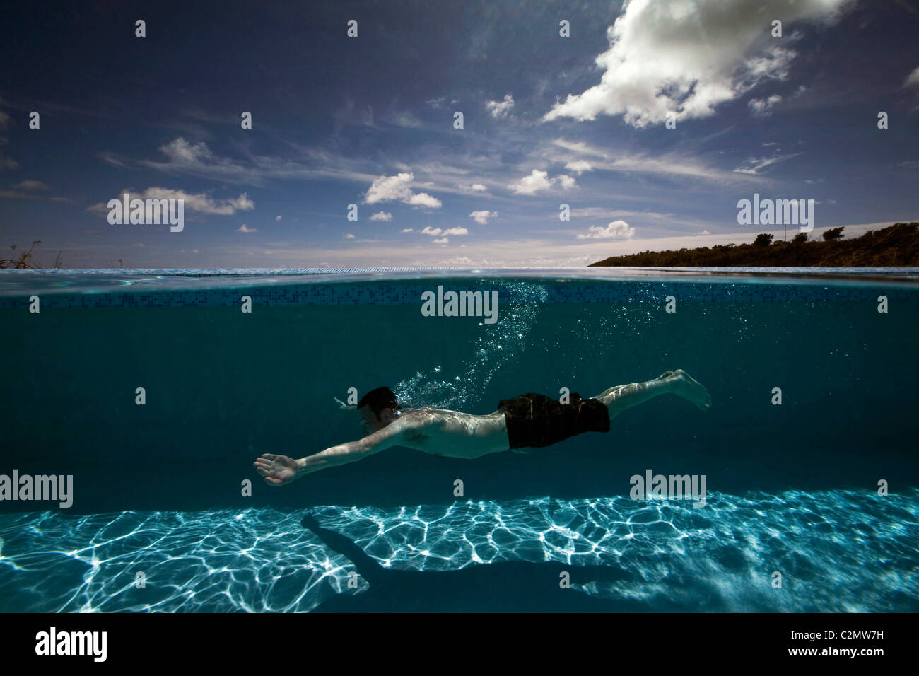 Un hombre nadando bajo el agua en una piscina de borde infinito bajo un cielo azul en un resort en la isla de Antigua en el Caribe. Foto de stock