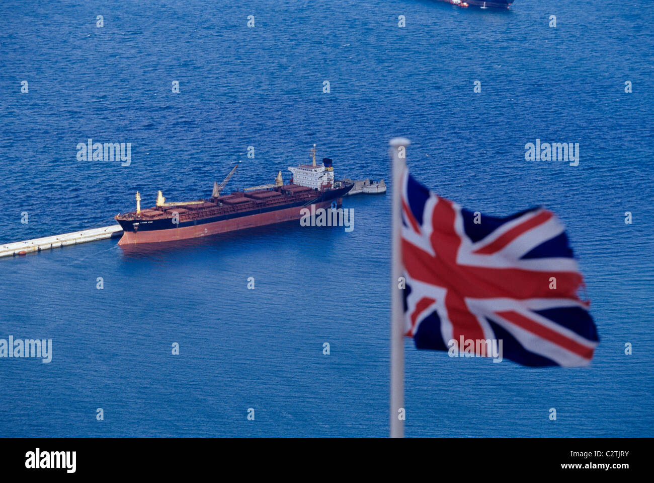 Una Union Jack bandera ondeando sobre el puerto de Gibraltar con un buque de carga en el fondo Foto de stock