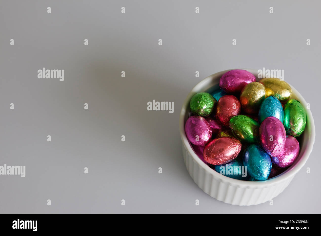 Un tazón de diminutos huevos de chocolate, envuelto en lámina de múltiples colores contra un fondo gris. Foto de stock