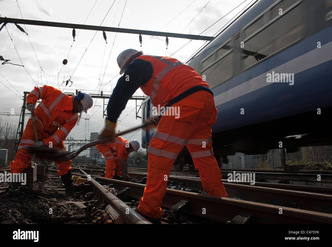 Los trabajadores de los ferrocarriles de ropa de alta visibilidad naranja  la reparación de una línea de ferrocarril de cercanías en Londres con el  tren en movimiento en segundo plano Fotografía de