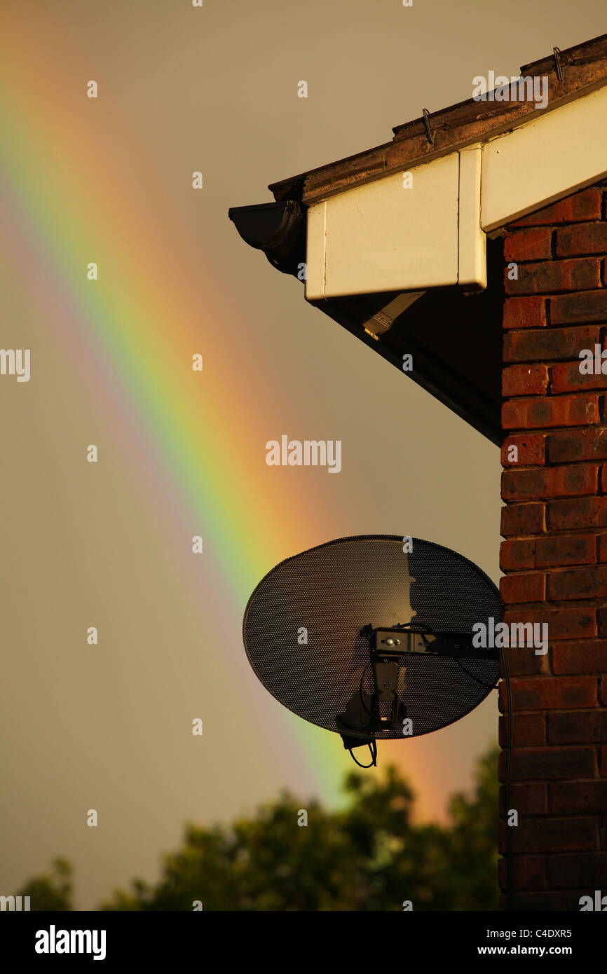 Un arco iris en un día tempestuoso con una casa y una antena parabólica en primer plano Foto de stock