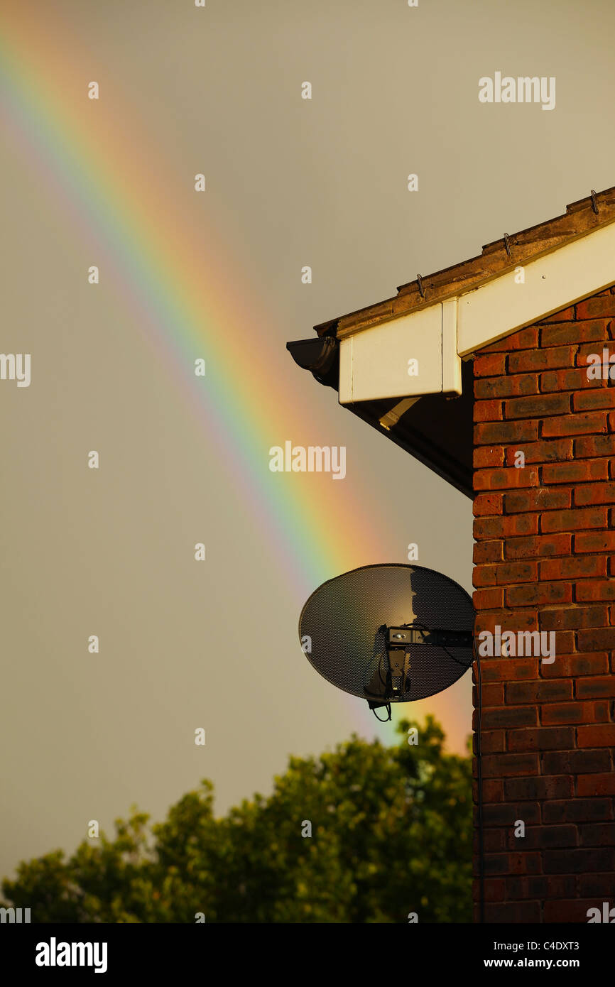 Un arco iris en un día tempestuoso con una casa y una antena parabólica en primer plano Foto de stock