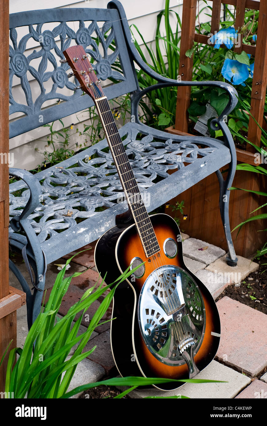 Un Dobro guitarra acústica con estilo sunburst finish se inclina contra un banco de trabajo de metal en un jardín. Foto de stock