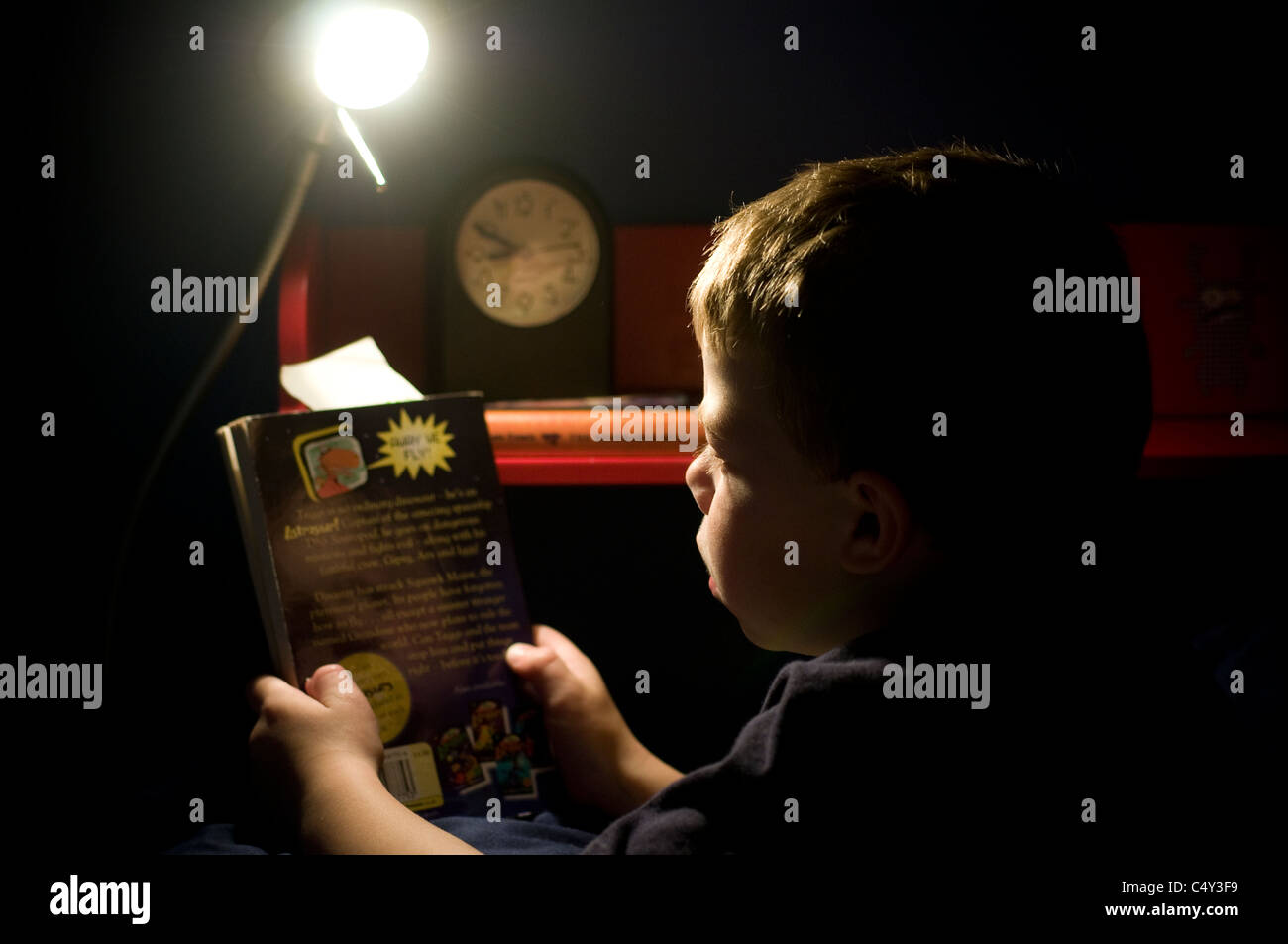 Reservar a la hora de acostarse, un niño de siete años leyendo un libro en la cama con una lámpara,chico leyendo en la noche, la familia, el ingenio para leer Foto de stock