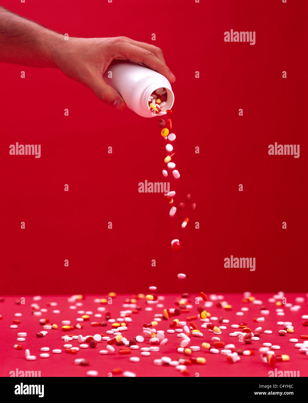 Una mano de hombre sosteniendo un contenedor mientras estudiando píldoras y tabletas sobre una superficie roja dispersos Foto de stock