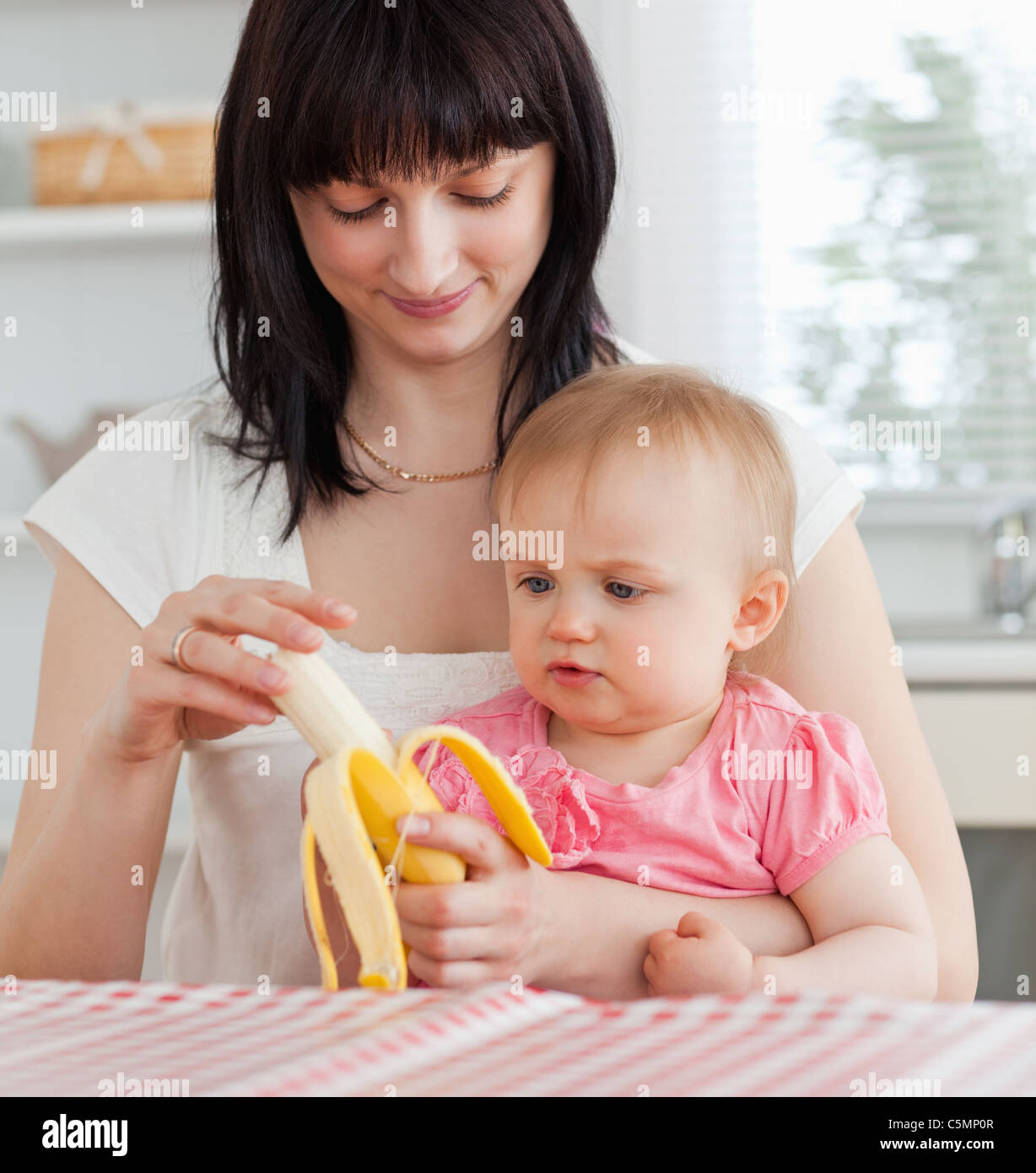 Encantadora mujer morena de pelar una banana mientras sostiene a su bebé Foto de stock