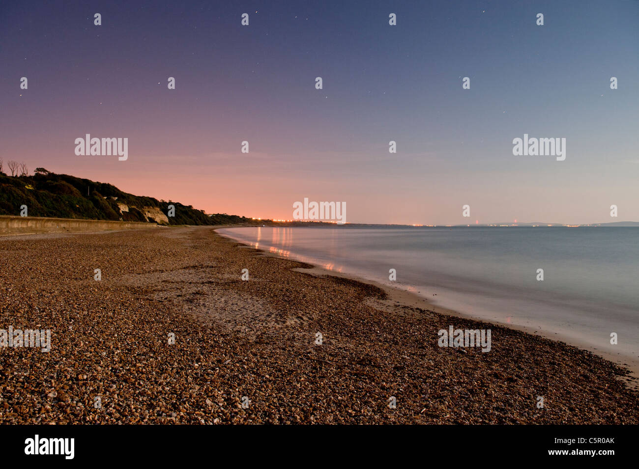 Luces Lejanas reflejan todavía un mar de noche por una playa de guijarros cerca de Highcliffe, Dorset. Foto de stock