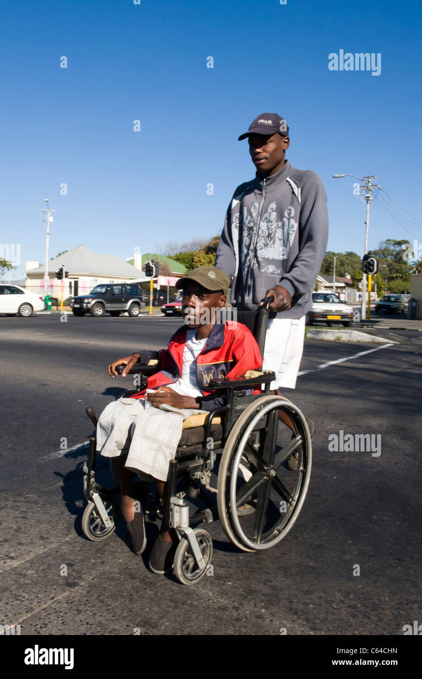Niño en una silla de ruedas es asistido por un cuidador que cruzar una carretera en Cape Town South Africa Foto de stock