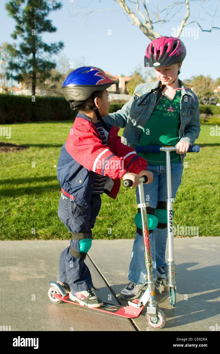 Vistiendo Chaqueta niña y niño hispano de pre-K en scooters en el parque  niño ayudando a otro señor © Myrleen Pearson Fotografía de stock - Alamy