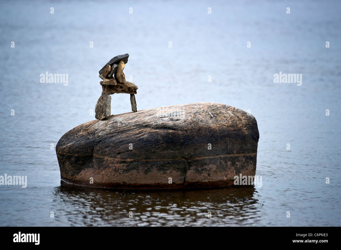 Un pequeño inuksuk (inuckshuk) se sitúa en la cima de una roca solitaria en el agua circundante. Foto de stock