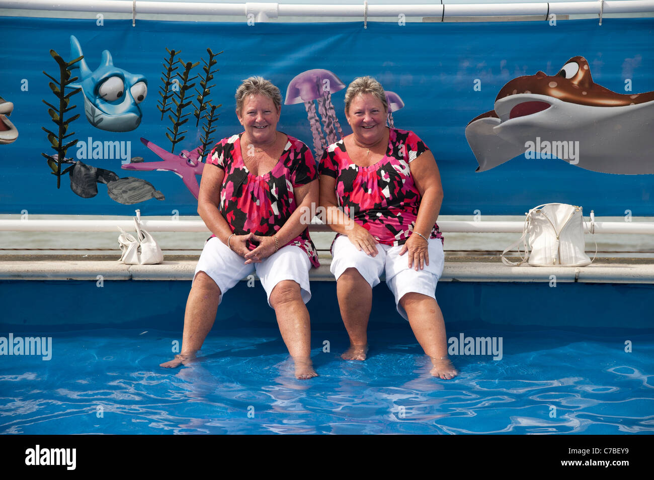 Dos hermanas gemelas idénticas de mediana edad vistiendo ropa coincidentes en la piscina Aberystwyth, Gales, Reino Unido, tarde de verano Foto de stock