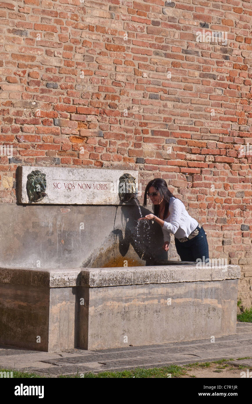 Una joven mujer adulta bebe de la fuente comunitaria junto al edificio público de Città della Pieve, Umbría, Italia. Foto de stock