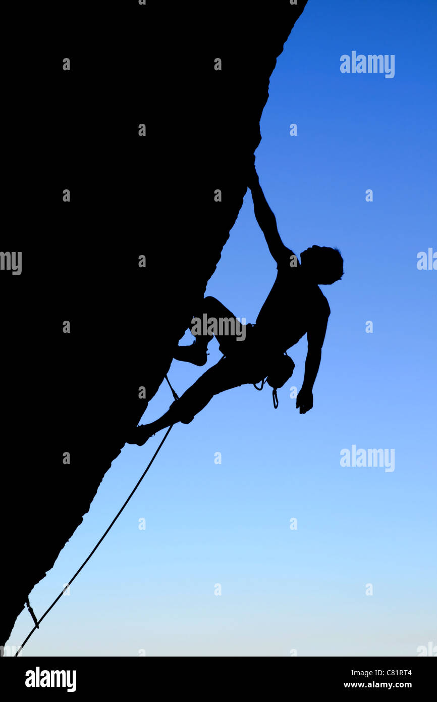 Silueta de escalador escalando un acantilado sobresaliente con fondo de cielo azul Foto de stock