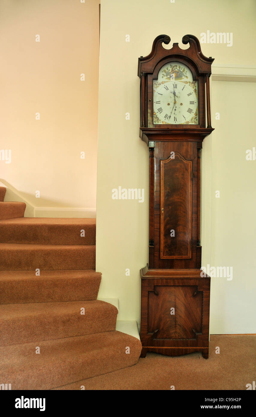 Un reloj de pie se encuentra en la base de la escalera en el pasillo de una casa. Foto de stock