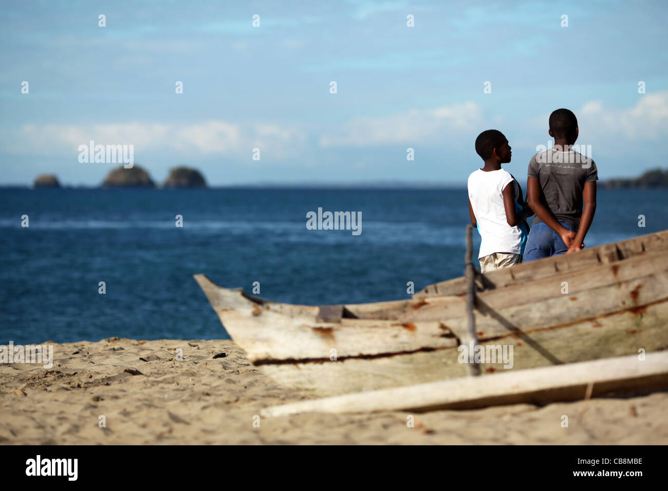 Dos jóvenes hombres contemplativos chat en la playa cercana a una canoa Outrigger malgache, con pequeñas islas en el fondo. Foto de stock