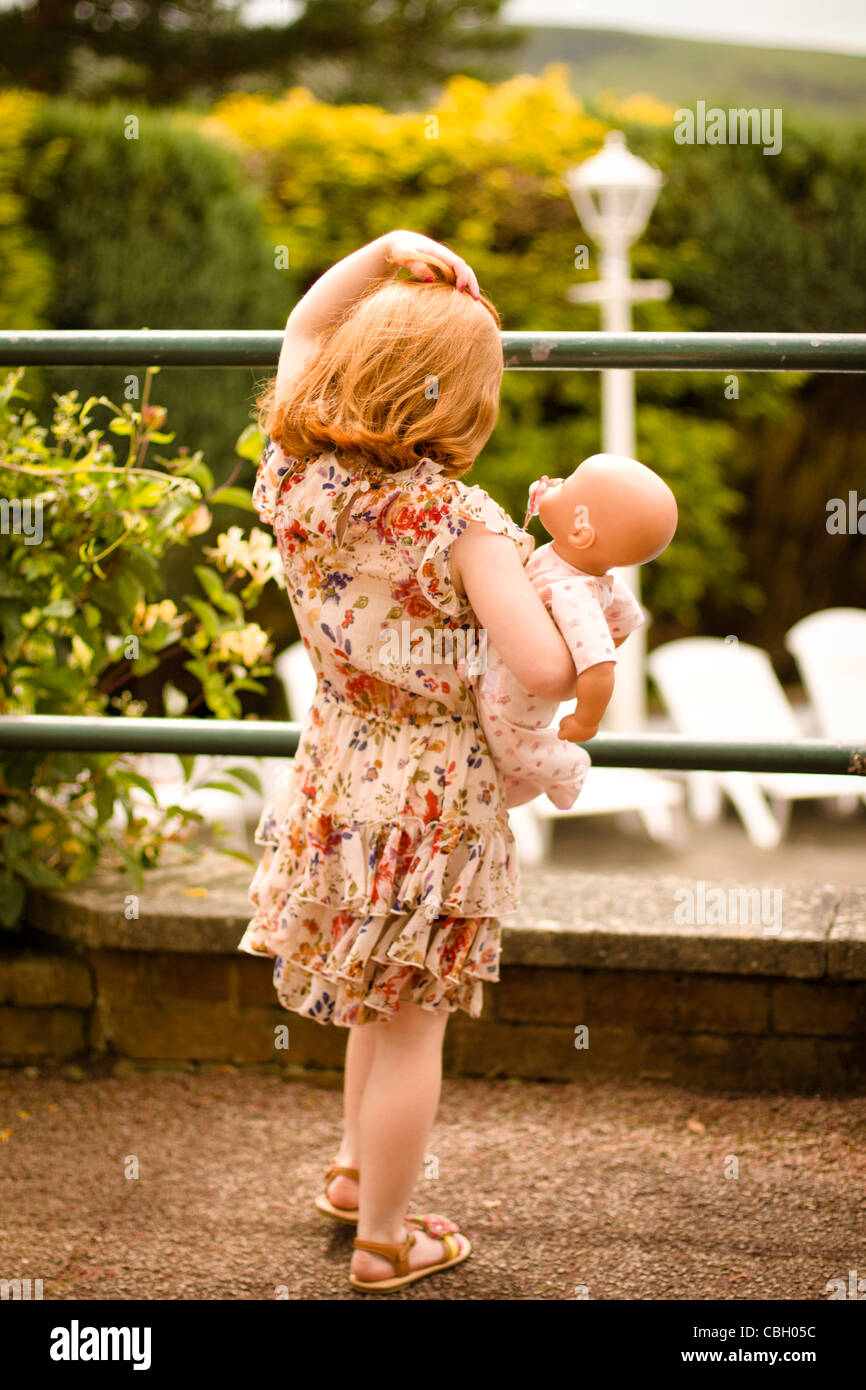 La parte trasera de una niña, con 5 años de edad, sosteniendo una muñeca, mirando a través de un jardín de verano Foto de stock