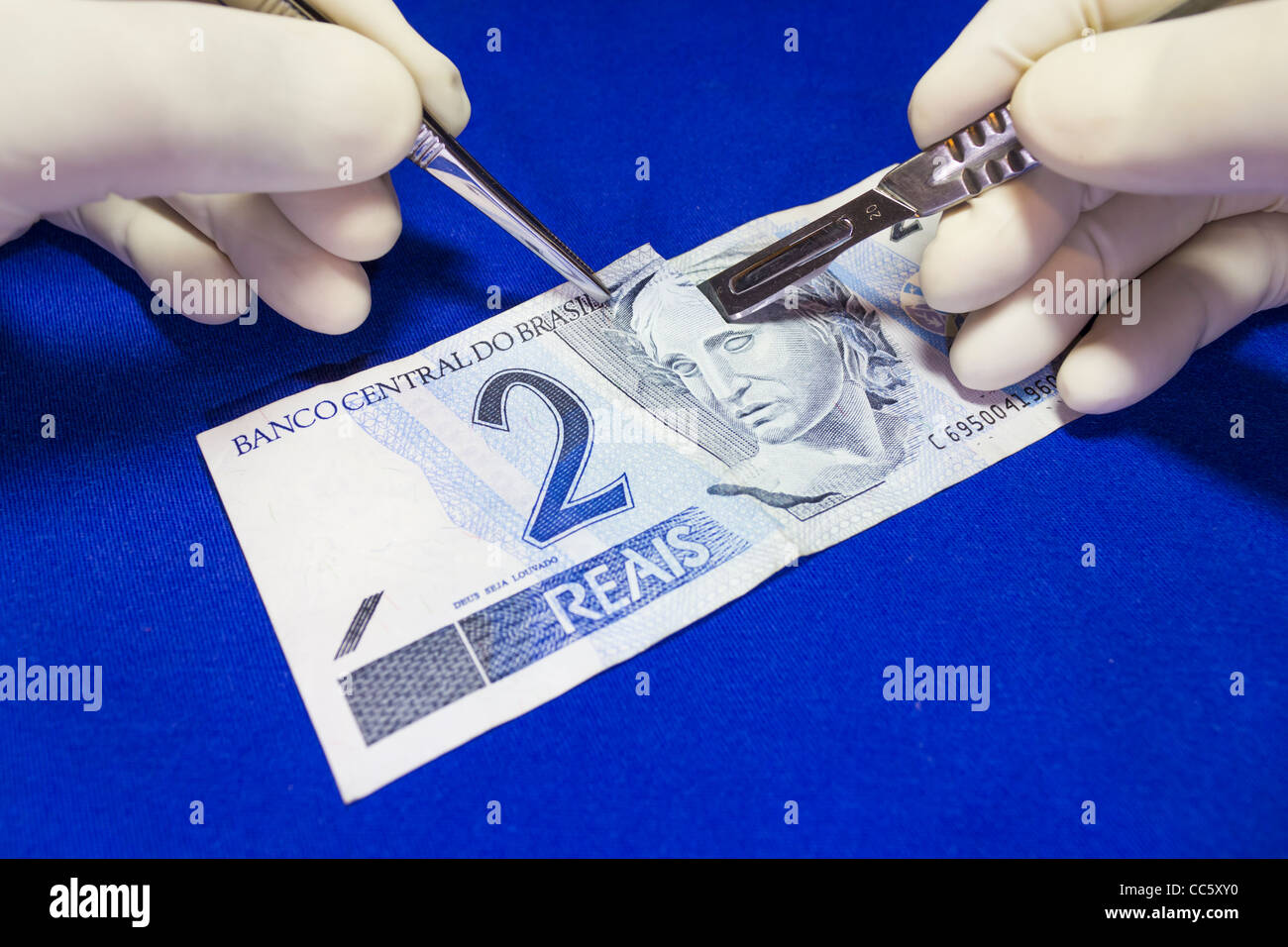 Cirugía - Dinero real brasileño. Un cirujano realiza una incisión quirúrgica en una nota con bisturí, pinzas y guantes estériles Foto de stock