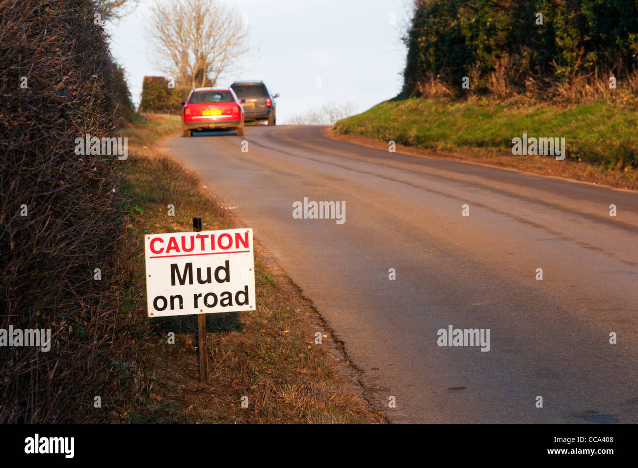 "Precaución barro en la carretera' firmar en un país de Norfolk lane con dos coches en la distancia. Foto de stock