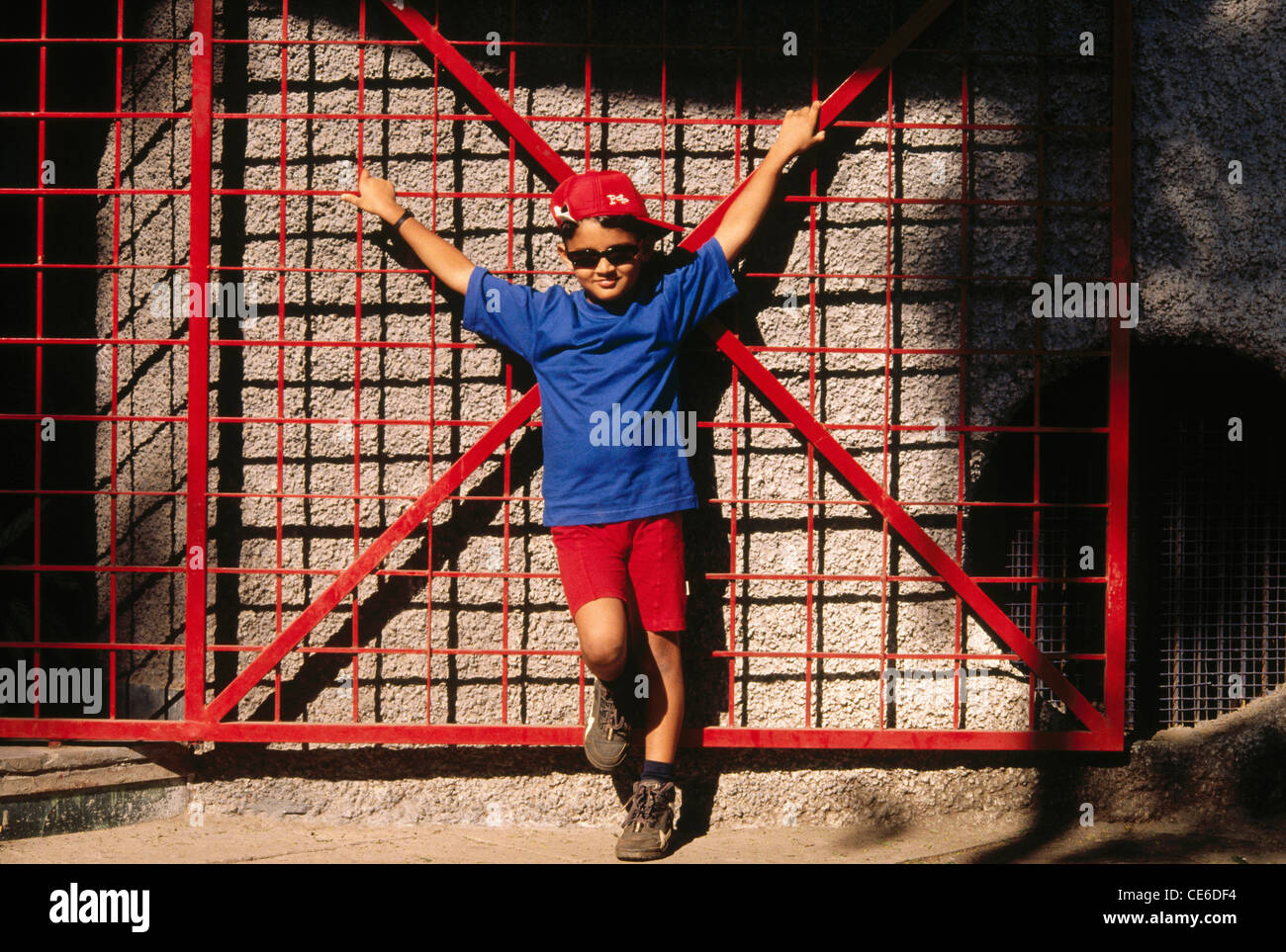 Chico en azul tshirt y calzones rojos y tapón rojo de pie con ambas manos  hacia arriba en la parte delantera del bastidor de metal rojo gate MR#158  Fotografía de stock -
