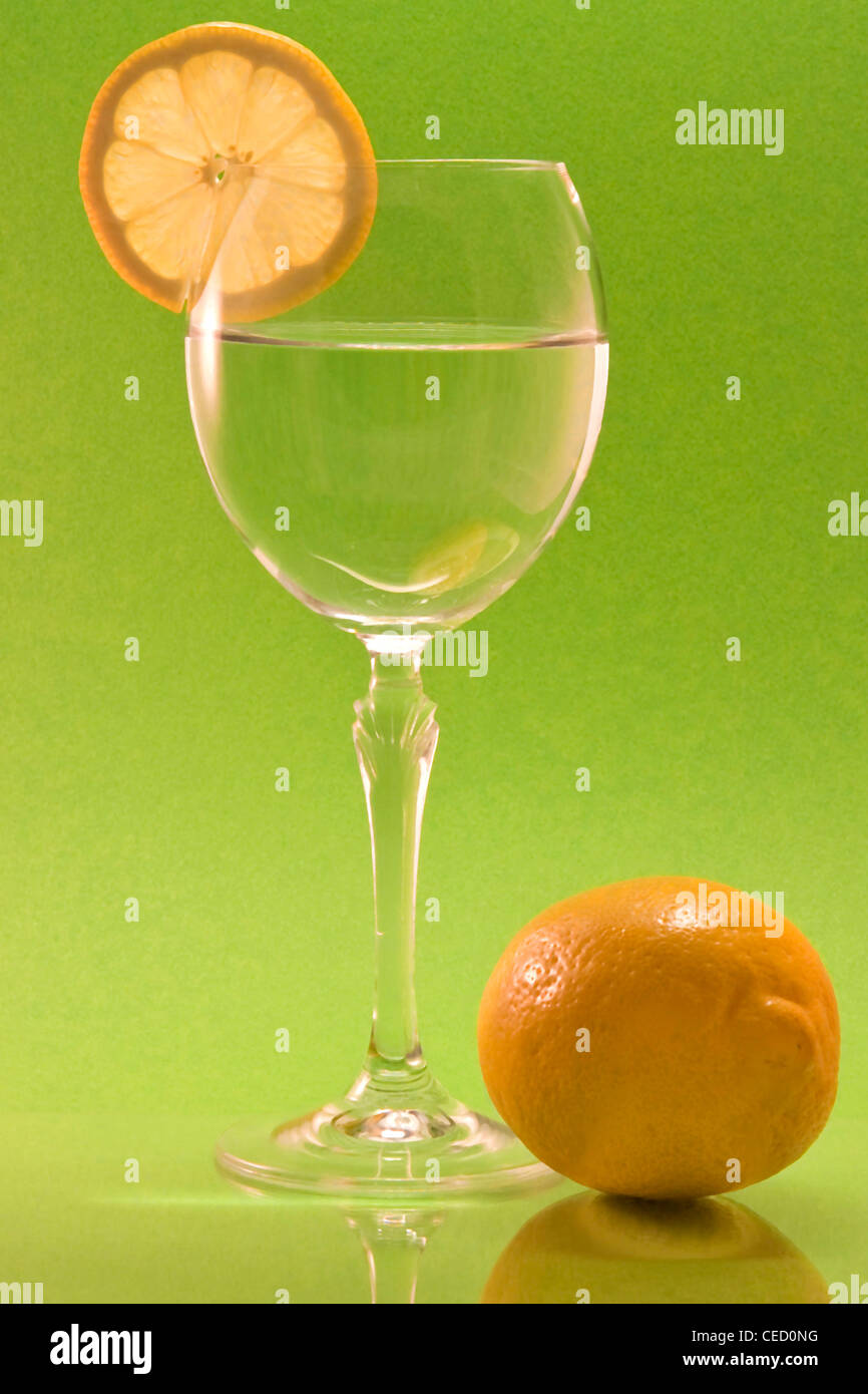 Una copa de vino con una rodaja de limón y un limón entero en la parte delantera y un fondo verde Foto de stock