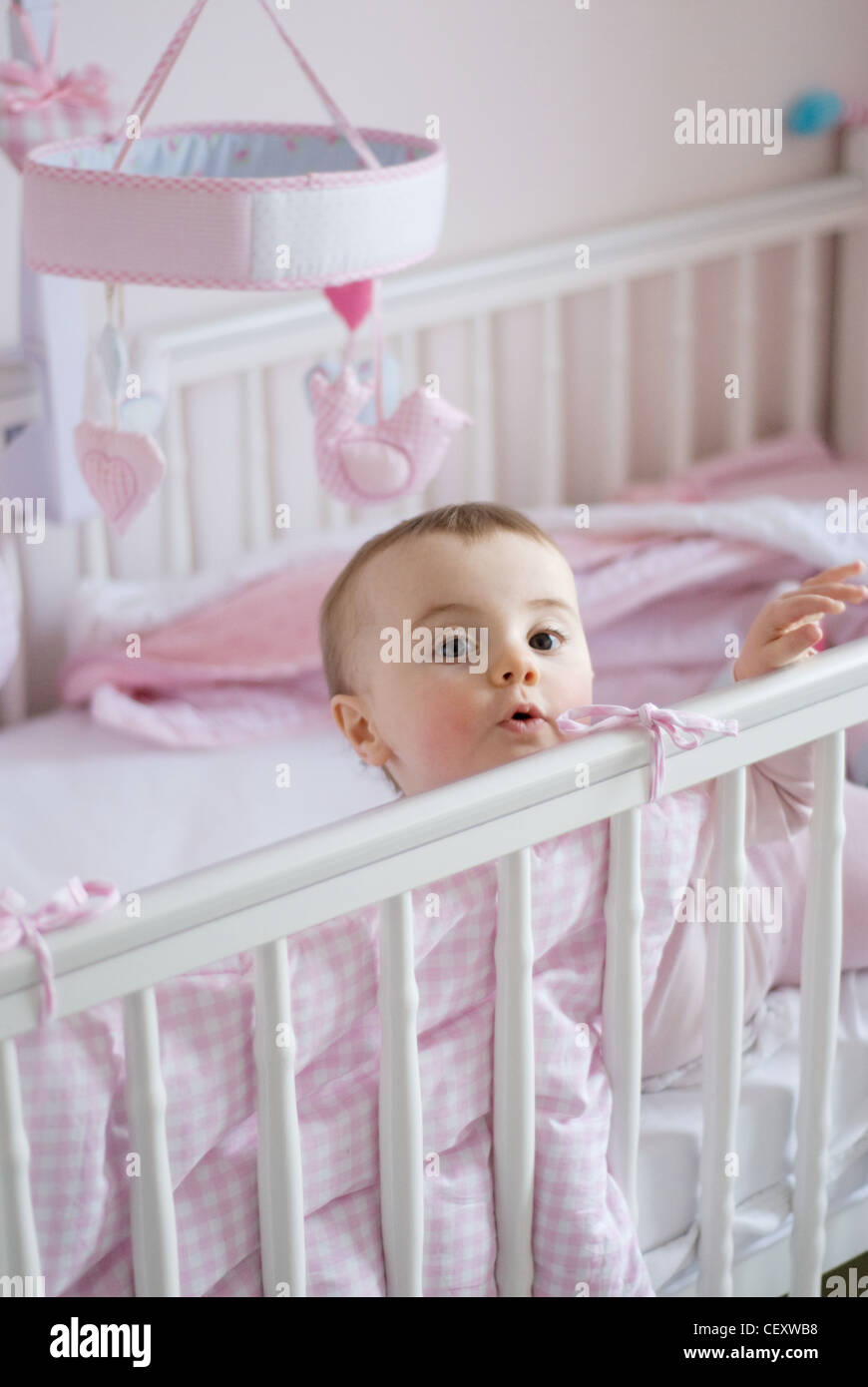 Un bebé hembra de cabello sentada en la cama cuna blanca rosa rosa y sábanas cuna móvil, nunca mirando a la cámara Penn Fotografía de stock -