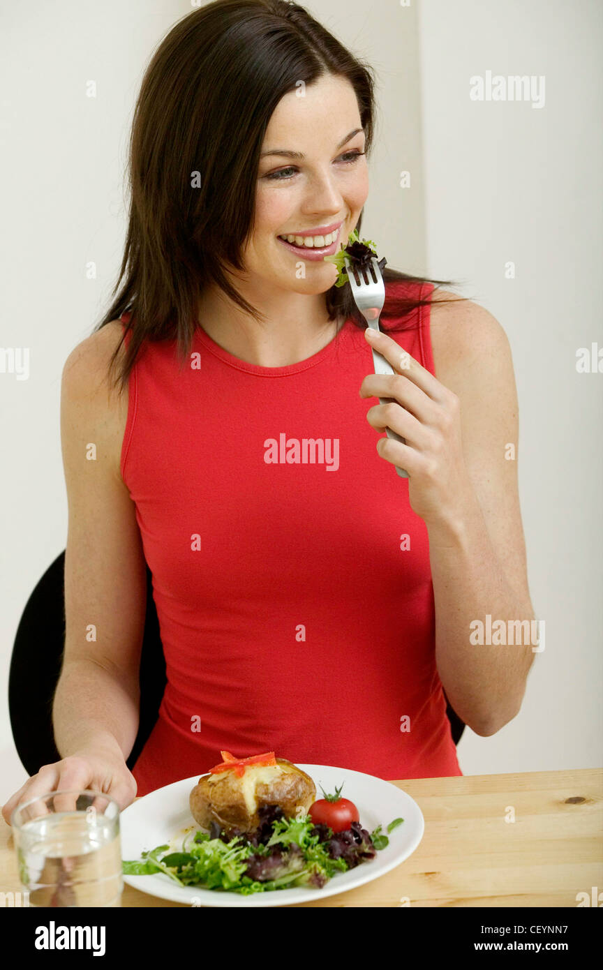 Mujer morena de largo cabello vestidos de rojo sin mangas sentados a la  mesa mantiene la horquilla a la lechuga, papas horneadas boca placa de  queso Fotografía de stock - Alamy