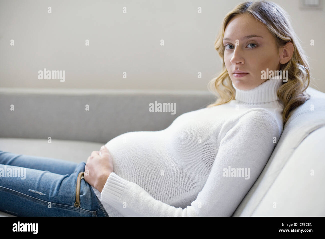 Una hembra preñada vistiendo pantalones azules blanco con cuello arriba, sentado en un sofá una manos sobre su estómago embarazada Fotografía de stock - Alamy