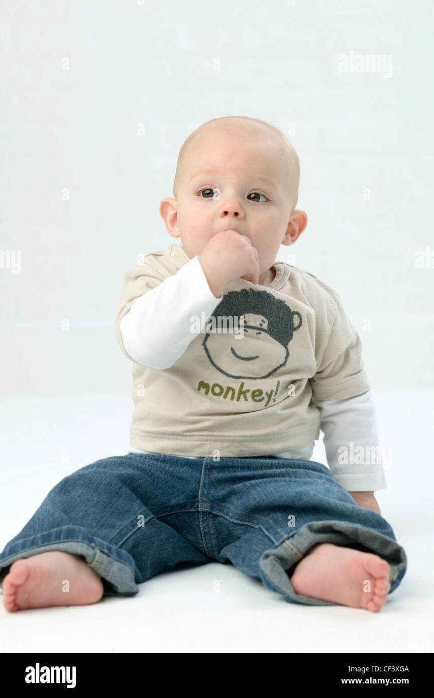 Bebé varón vistiendo mono beige camiseta, top blanco y azul denim jeans,  sentado en flowith mano a boca, contra un blanco Fotografía de stock - Alamy